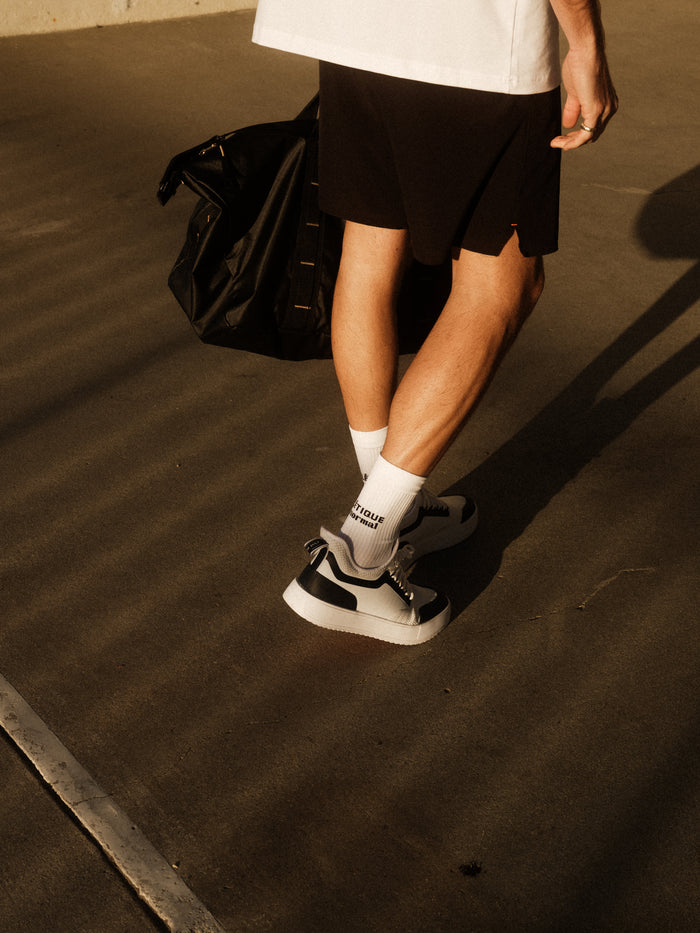 A person wearing white The Athlétique Socks, black shorts, and sneakers stands outdoors on concrete, carrying a black duffel bag. The image focuses on the lower legs, highlighting the black c'est normal logo on the mid-calf of the socks.