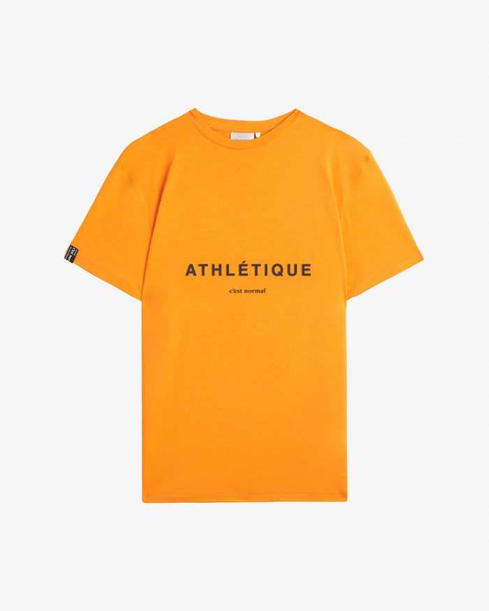 A front view of The Athlétique Tee in orange, laid flat on a white background. The image highlights the clean crewneck, the Athlétique and c'est normal logo printed on the chest, and a small tag on the right sleeve.