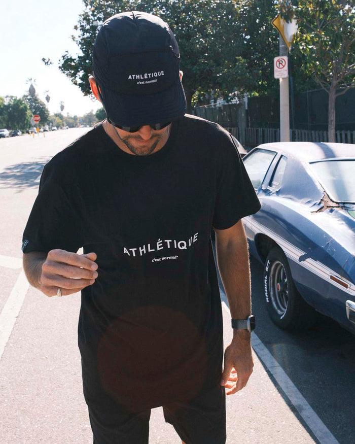 A man stands outdoors wearing The Athlétique Tee in black, matching shorts, sunglasses, and a black cap. The image highlights the relaxed fit of the t-shirt and the white Athlétique logo with c'est normal written underneath on both the cap and chest.