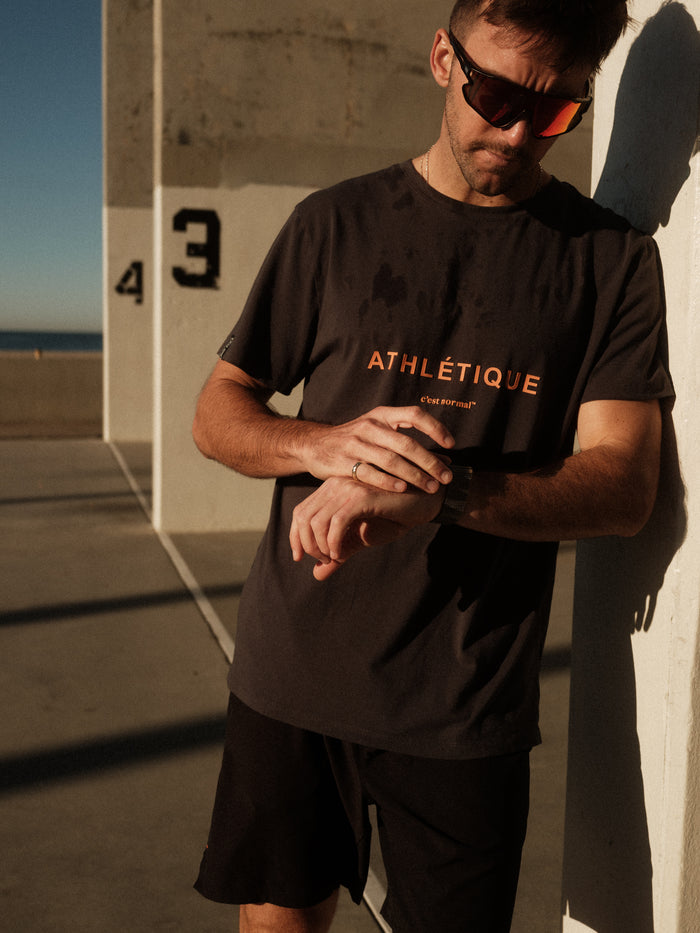 A man wearing The Athlétique Tee in asphalt and sunglasses leans against a white outdoor pillar. He is looking down at his watch, highlighting the printed ATHLÉTIQUE and c'est normal logos on the chest.