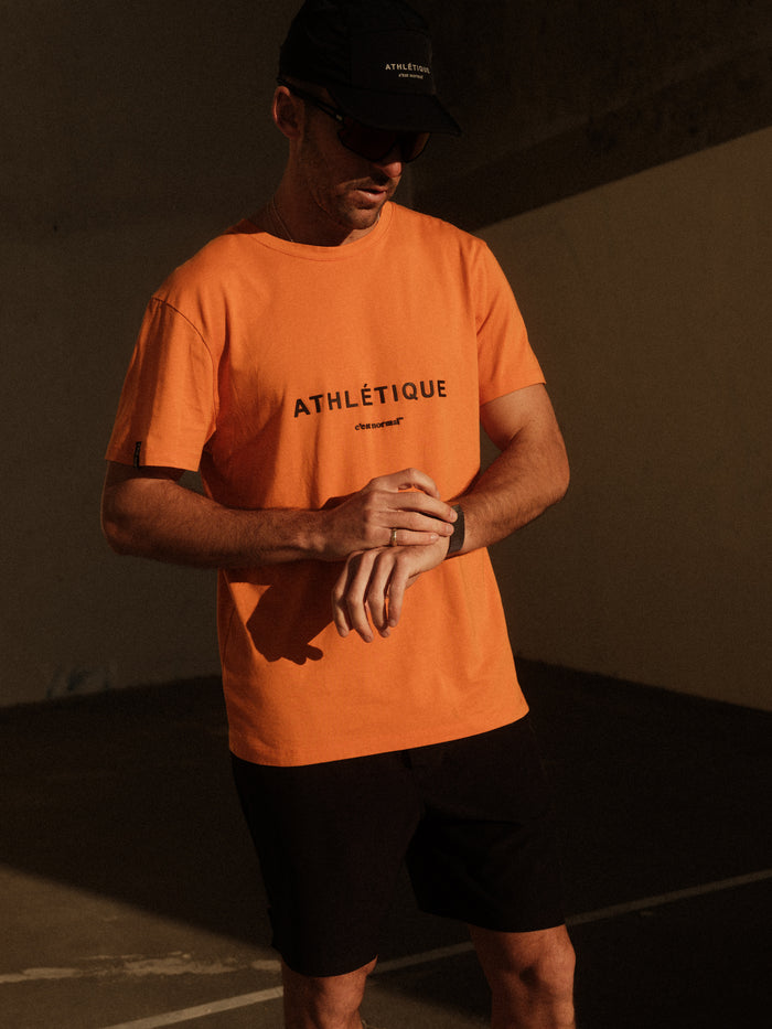 A man wearing The Athlétique Tee in orange, featuring the ATHLÉTIQUE logo and c'est normal text on the chest. He is adjusting his watch while standing in the sunlight, paired with a black cap and black shorts.