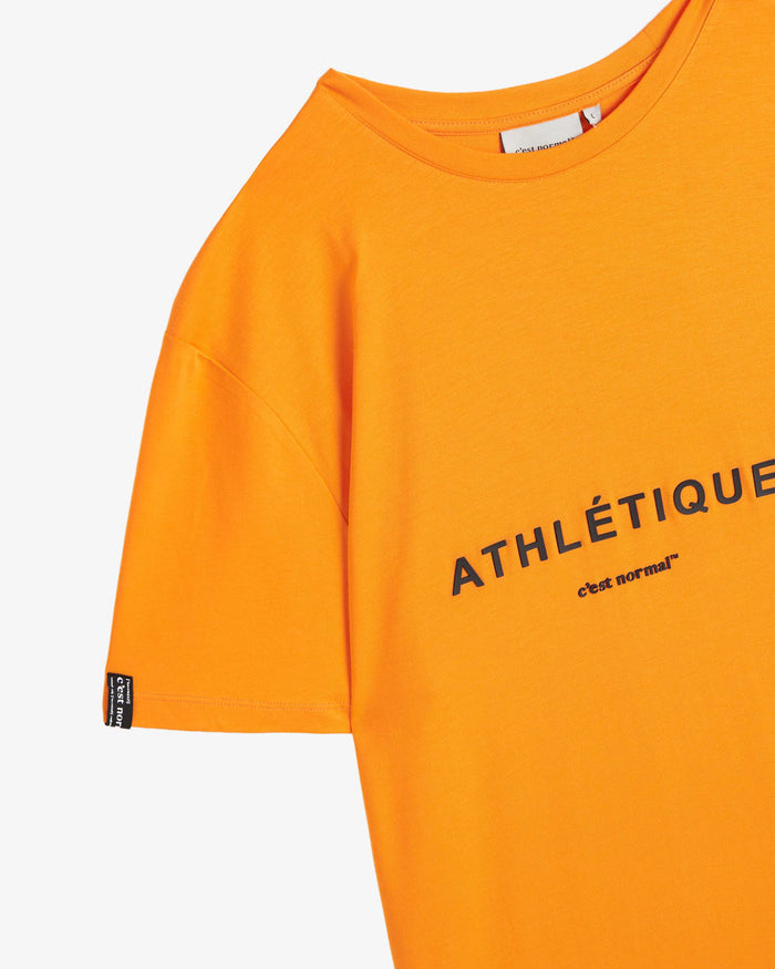 A close-up of The Athlétique Tee in orange, focusing on the crewneck, the black cuff tag on the sleeve, and the printed Athlétique and c'est normal logo on the chest.