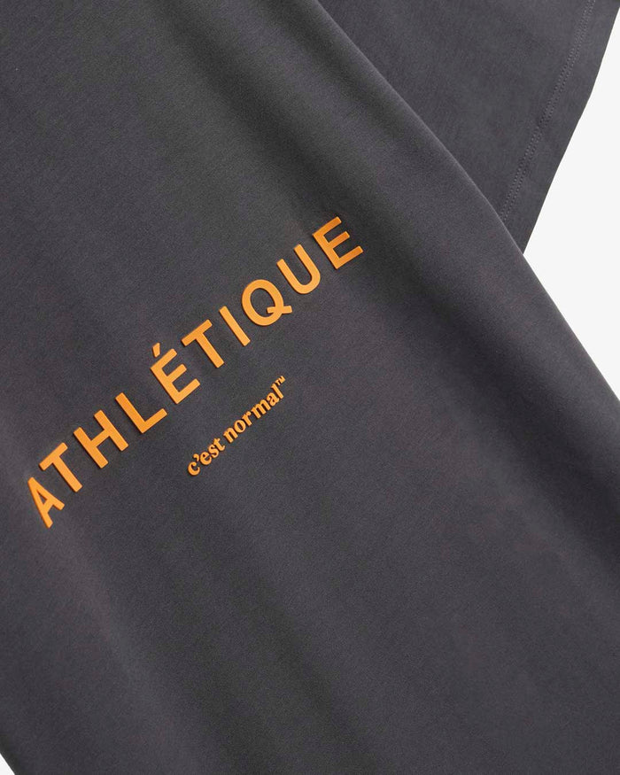 A detailed close-up of The Athlétique Tee in asphalt grey, showing the orange ATHLÉTIQUE print and the small c'est normal logo on the soft, natural fiber blend fabric.