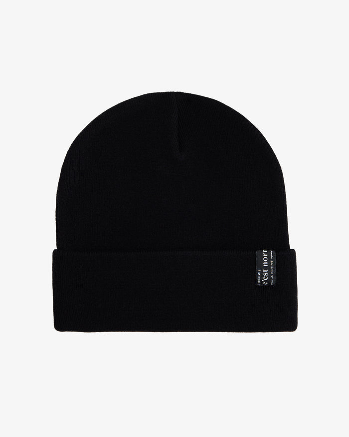 A front view of The Beanie in black, laid flat on a white background. The image highlights the fold-over cuff and the signature c'est normal label.