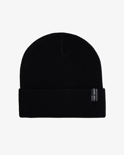 A front view of The Beanie in black, laid flat on a white background. The image highlights the fold-over cuff and the signature c'est normal label.