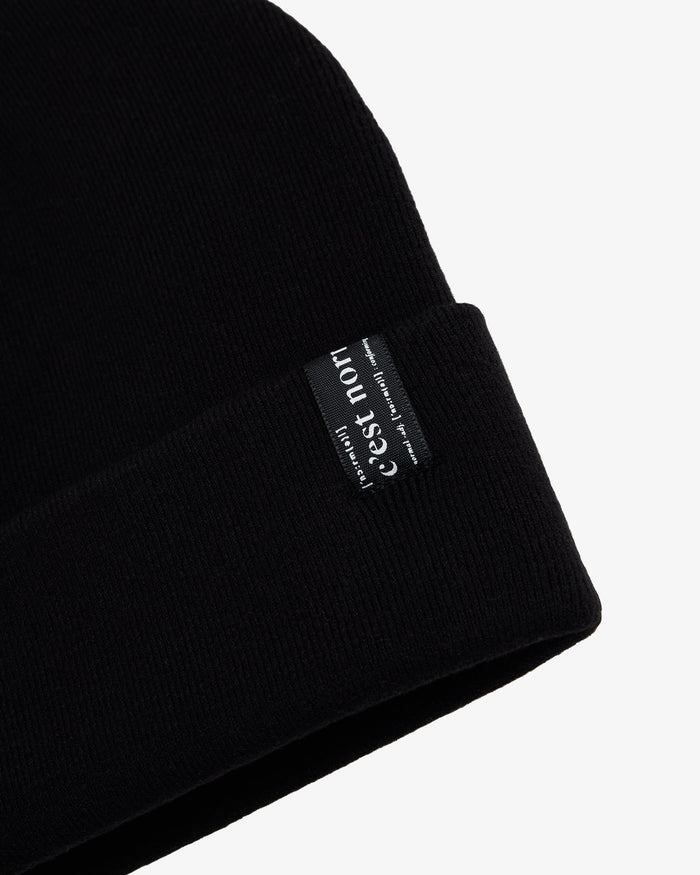 A close-up shot of The Beanie in black, laid flat on a white background, focusing on the fold-over cuff and the signature c'est normal label.