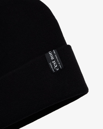 A close-up shot of The Beanie in black, laid flat on a white background, focusing on the fold-over cuff and the signature c'est normal label.