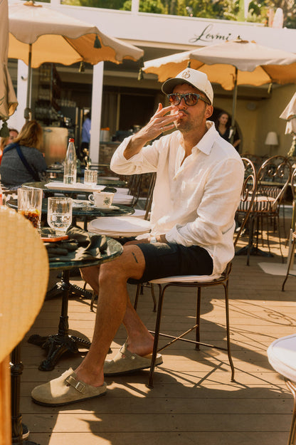 The Board Shorts in black — worn with a white linen shirt and cap while sitting at a sunlit cafe