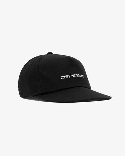 A front view of The Caddy Cap in black, laid flat on a white background. The image highlights the solid black color, flat visor, and the embroidered c'est normal logo on the front.