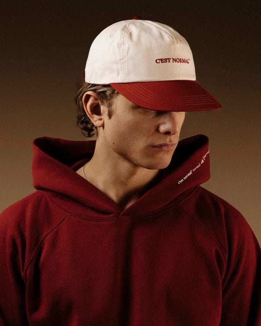 A man wearing The Caddy Cap in cream and crimson, paired with a matching crimson hoodie. The image highlights the cap's cream crown, crimson visor, and the red embroidered c'est normal logo on the front.
