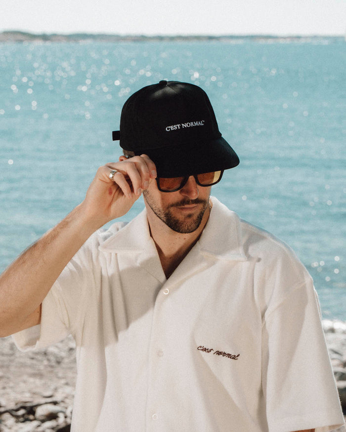 A man wearing The Caddy Cap in black, sunglasses, and a white short-sleeve shirt, stands in front of the sparkling ocean. He is adjusting the brim of the cap, which features the embroidered white c'est normal logo.