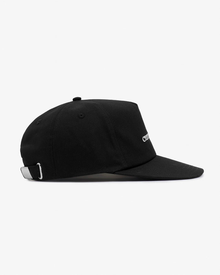 A side view of The Caddy Cap in black against a white background. The image highlights the cap's silhouette, the adjustable strap with a metal buckle at the back, and the partially visible embroidered c'est normal logo on the front.