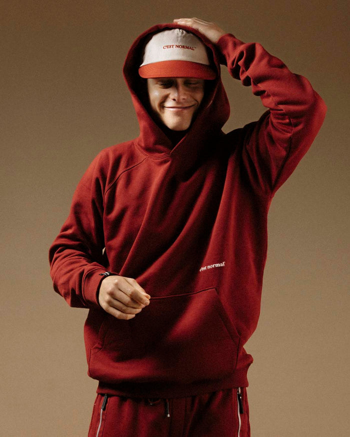 A smiling young man wearing The Caddy Cap in Cream/Crimson and a red hoodie with the hood up. The cap has a cream top with the c'est normal logo embroidered in red, and a crimson visor.