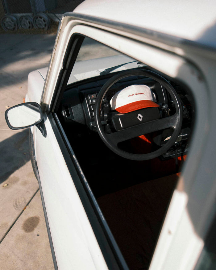 A view through the open window of a white car, showing The Caddy Cap in Cream/Crimson resting on the steering wheel. The cap features a cream crown, a crimson visor, and the embroidered c'est normal logo on the front.