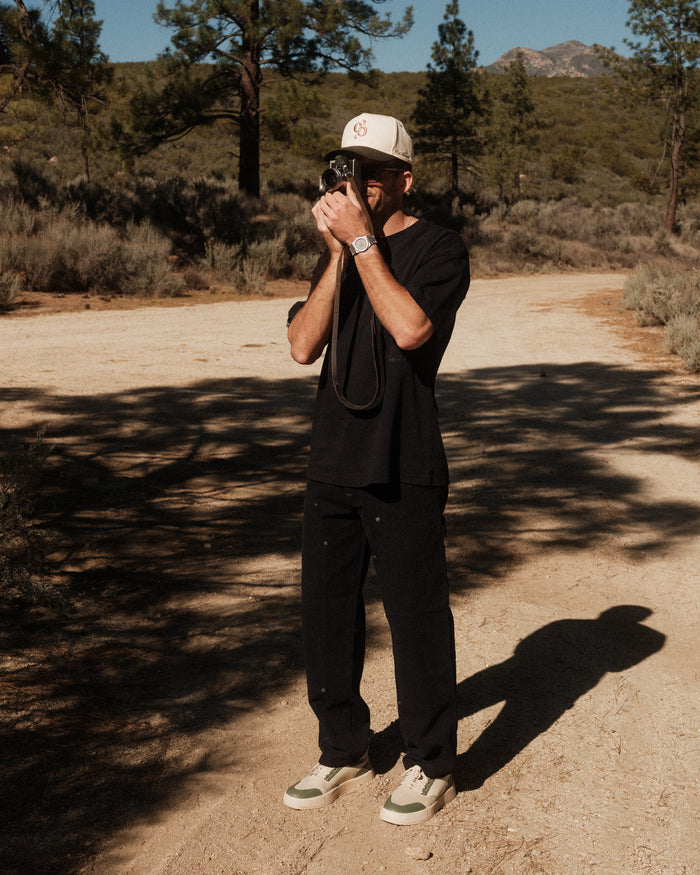 A man wearing The Carpenter Pant in black, a black t-shirt, and a c'est normal cap stands on a dirt road, holding a vintage camera up to his face. The background features a landscape with trees and distant hills.