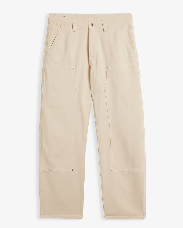 A front view of The Carpenter Pant in sand, laid flat on a white background. The image highlights the double-knee panels, copper-toned rivets, and the embroidered c'est normal logo by the left pocket.