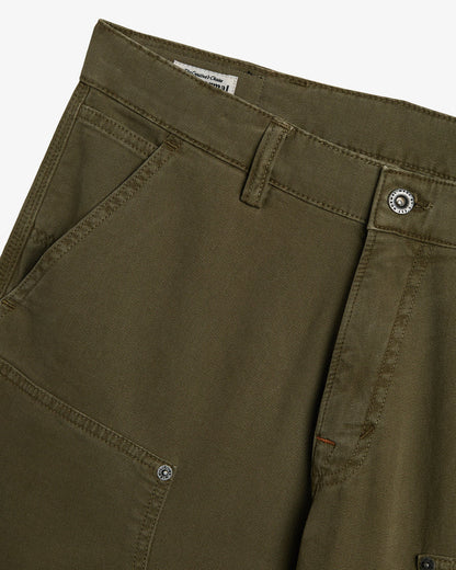 A close-up shot of the front of The Carpenter Pant in olive green, showcasing the textured fabric, a slash pocket, and custom c'est normal buttons and rivets.