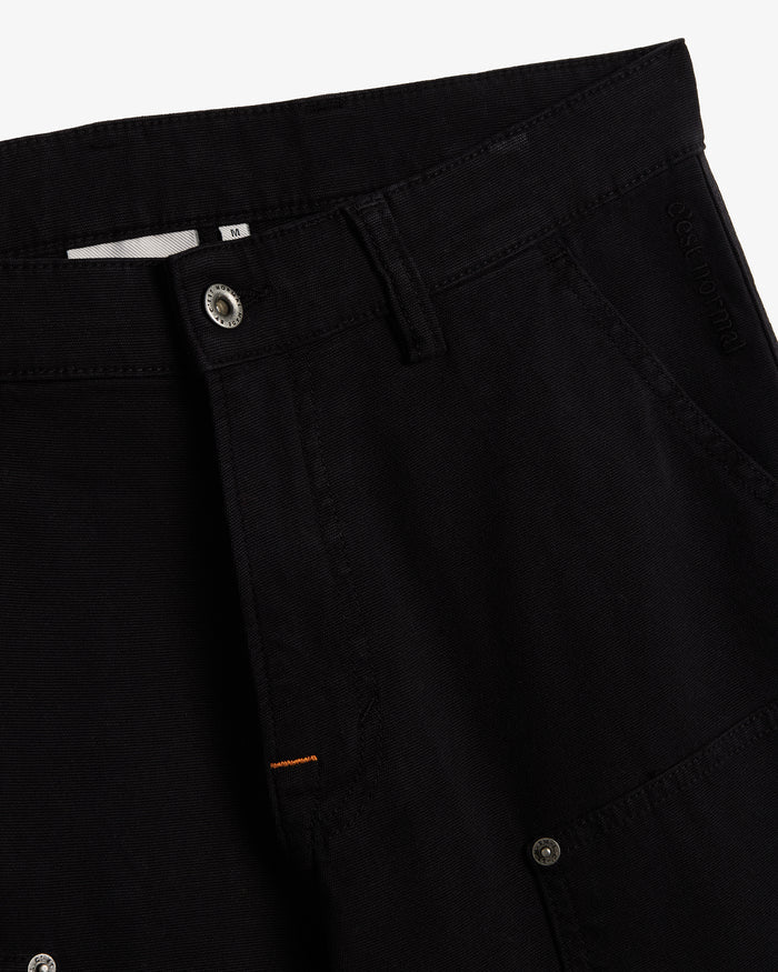 A detailed close-up of The Carpenter Pant in black, highlighting the front button closure, textured fabric, and the embroidered c'est normal logo near the pocket.