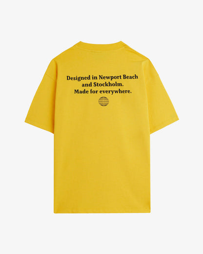 A back view of The c'est pari Oversized T-Shirt in yellow, laid flat on a white background. The image highlights the black screen printed text on the back that says "Designed in Newport Beach and Stockholm. Made for everywhere." with a globe icon below it.