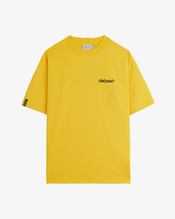 A front view of The c'est pari Oversized T-Shirt in yellow, laid flat on a white background. The image highlights the c'est pari logo embroidered on the chest and a c'est normal cuff tag on the right sleeve.