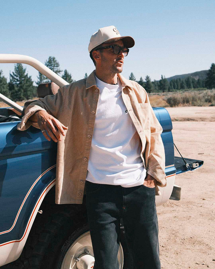 A man leaning against a blue pickup truck outdoors, wearing The CÔTELÉ Corduroy Overshirt in Irish Cream over a white t-shirt and black pants, along with a two-tone c'est normal cap and sunglasses.