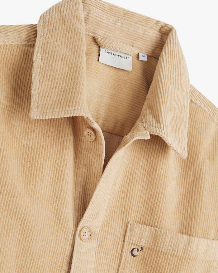 A close-up of The CÔTELÉ Corduroy Overshirt in Irish Cream, focusing on the collar, front buttons, and chest pocket. The image highlights the ribbed corduroy texture and the inner c'est normal label.