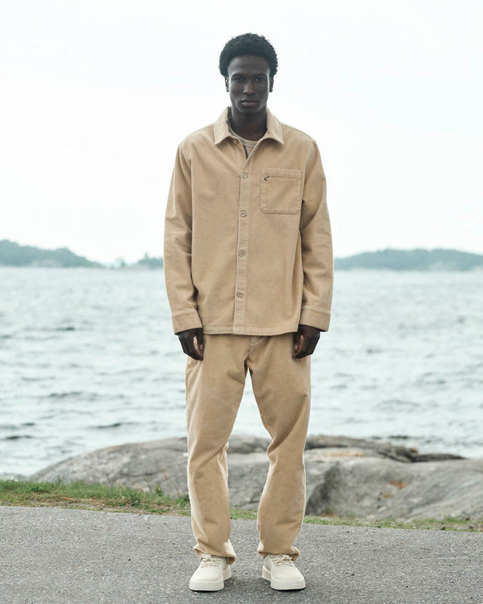 A model standing outdoors near a body of water wearing The CÔTELÉ Corduroy Overshirt in Irish Cream, paired with matching corduroy pants. The overshirt features a straight fit, a front pocket with a small c'est normal logo, and button closures.