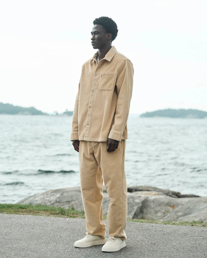 A man standing outdoors by the water, wearing The CÔTELÉ Corduroy Overshirt in Irish Cream, paired with matching corduroy pants and white sneakers.