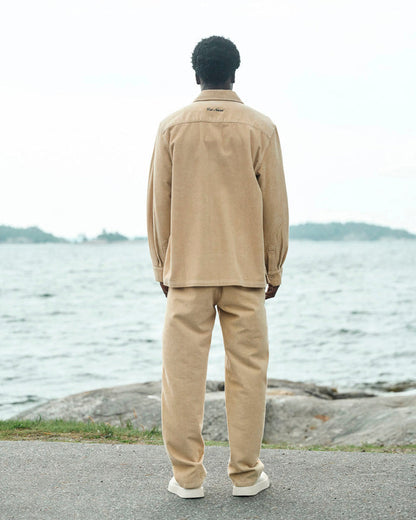 A back view of a man standing by the water wearing The CÔTELÉ Corduroy Overshirt and matching pants in Irish Cream. The image highlights the straight fit and the embroidered c'est normal logo at the back of the neck.