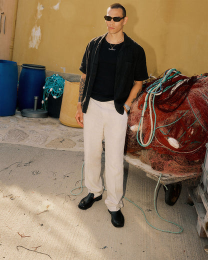 The Daily Linen Trouser in natural — worn with a black shirt and loafers near fishing nets