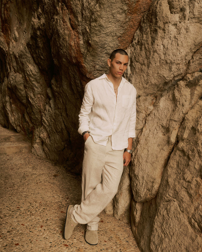 The Daily Linen Shirt in off-white — leaning against a rocky cliff wall