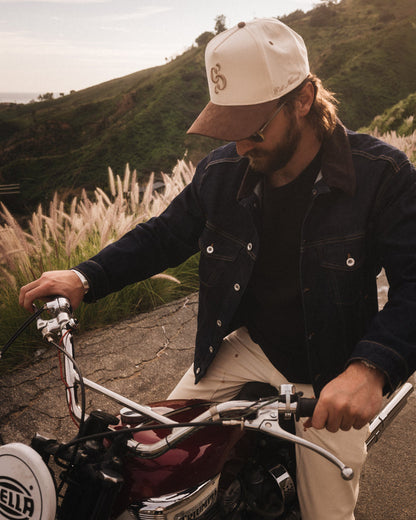 A man wearing The Snapback Cap in cream and brown, sitting on a motorcycle in a hilly landscape. The image highlights the c'est normal mirror logo embroidered on the front of the cap.