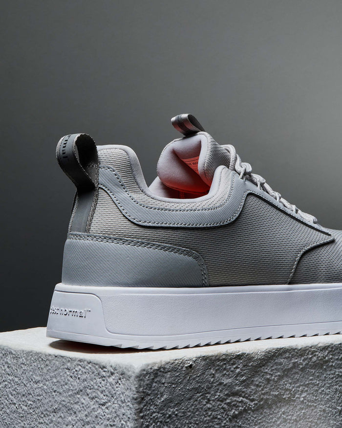 A detailed close-up of the heel of The Dynamic Waterproof Sneaker 2.0 Low in grey, focusing on the thick white sole with the embossed c'est normal logo and the heel pull tab. The image also showcases the padded collar and textured grey waterproof upper.