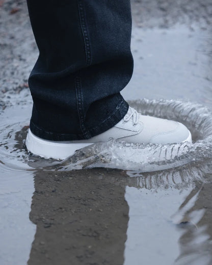 A close-up of a person wearing dark jeans and The Dynamic Waterproof Sneaker 2.0 Low in grey stepping into a puddle. The splashing water highlights the waterproof design of the c'est normal sneaker.
