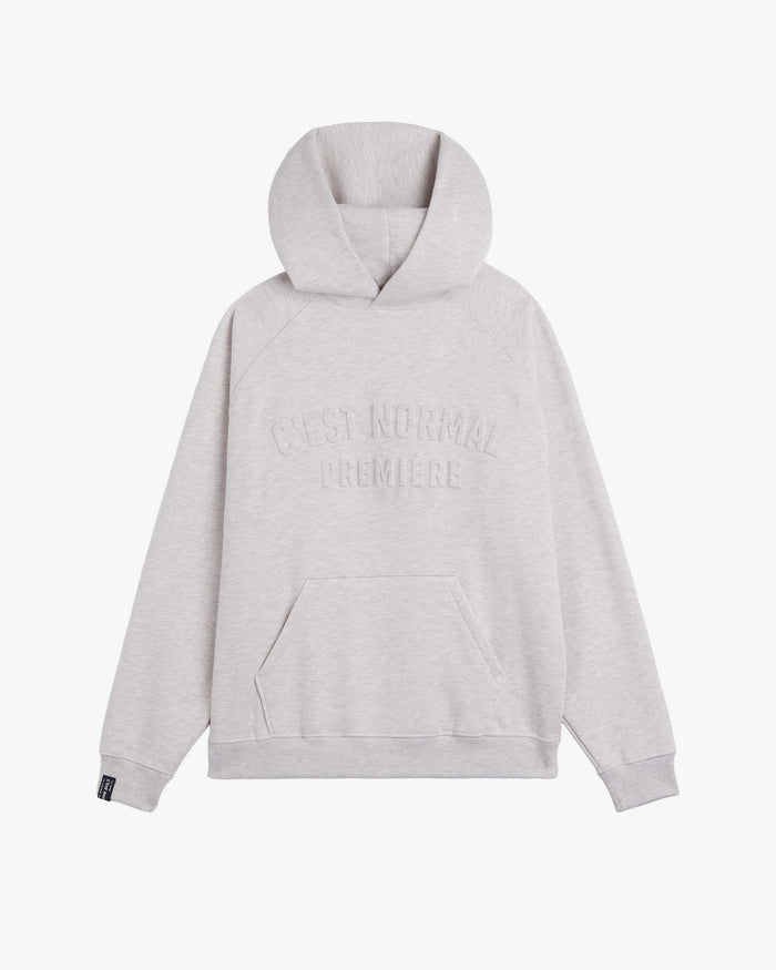 A front view of The Embossed Hoodie in beige melange, laid flat on a white background. The image highlights the embossed c'est normal logo on the chest, the kangaroo front pocket, and the ribbed cuffs and hem.