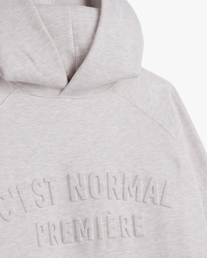 A close-up shot of The Embossed Hoodie in beige melange, highlighting the thick hood and the embossed c'est normal première logo on the chest.