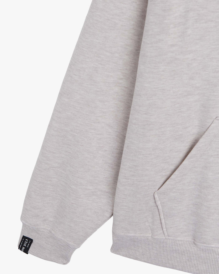 A detailed close-up of The Embossed Hoodie in beige melange, highlighting the right sleeve's ribbed cuff with the black c'est normal tag, and the edge of the kangaroo front pocket.