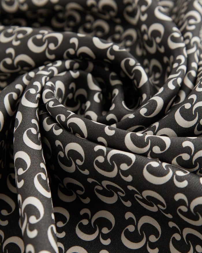 A close-up view of the lightweight woven fabric of The Fall Bandana, showcasing the signature repeating c'est normal monogram print in black and cream.