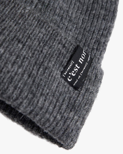 A detailed close-up of The Hairy Beanie in charcoal grey, focusing on the soft knitted texture and the black brim tag with the white c'est normal logo.