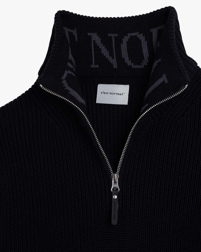 A detailed close-up of The Halfzip Sweater in black, focusing on the metal zipper and the black rubber puller with the c'est normal logo. The image also showcases the structured knit texture and the intarsia c'est normal logo inside the standing collar.