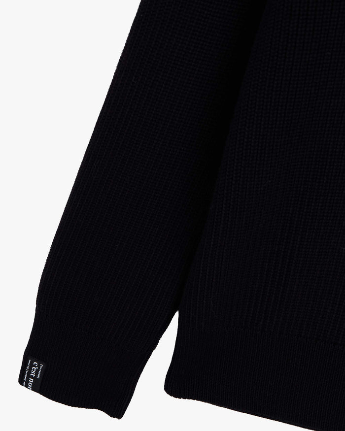 A close-up shot of the right sleeve of The Halfzip Sweater in black, laid flat on a white background. The image highlights the knit texture of the 100% cotton fabric and the black cuff tag with the white c'est normal logo.