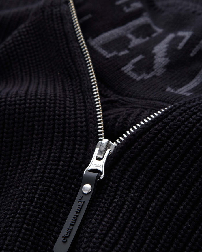 A detailed close-up of The Halfzip Sweater in black, focusing on the metal zipper and the black rubber puller with the c'est normal logo. The image also showcases the textured cotton knit material and the intarsia logo inside the collar.