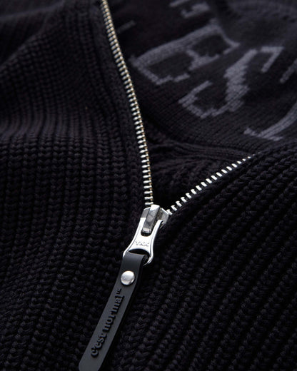 A detailed close-up of The Halfzip Sweater in black, focusing on the metal zipper and the black rubber puller with the c'est normal logo. The image also showcases the textured cotton knit material and the intarsia logo inside the collar.
