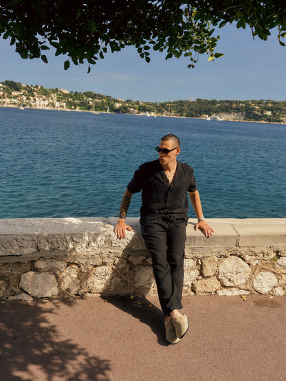The Holiday Linen Trouser in black — worn leaning against a stone wall by the sea