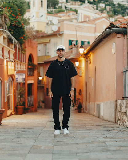A man stands in a cobbled street wearing The Jeans Relaxed Straight Fit in Jet Black, paired with a black t-shirt, white sneakers, and a white c'est normal cap.