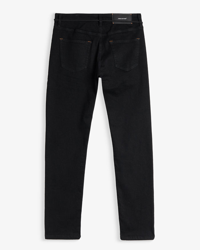 A back view of The Jeans Slim Fit in Jet Black, laid flat on a white background. The image highlights the two back pockets, subtle orange accent stitching, and a rubber patch at the back waist featuring the c'est normal logo.