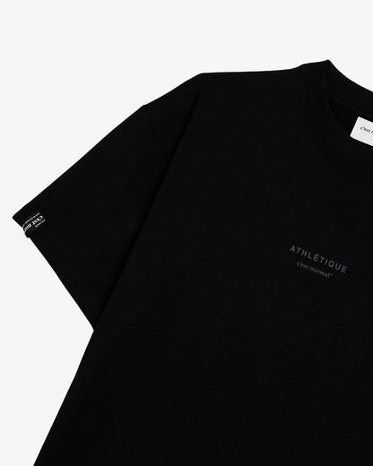 A close-up view of The Oversized Gym T-Shirt in black, laid flat on a white background. The image highlights the dark Athlétique logo on the front and the signature cuff tag with the c'est normal logo on the sleeve.