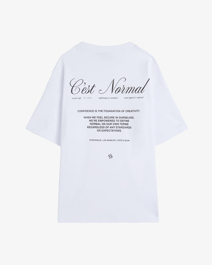 A back view of The Première T-Shirt in white, laid flat on a white background. The image highlights the signature c'est normal artwork and text printed in black on the back.