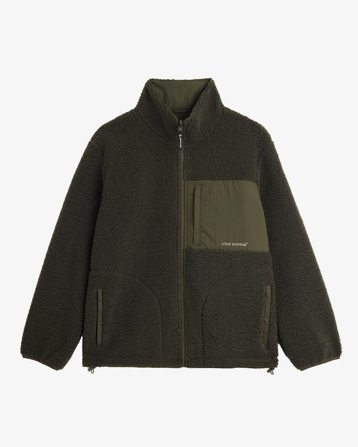A front view of The Reversible Pile Jacket in Forest Green, laid flat on a white background. The image highlights the cozy pile fleece side, the zippered chest pocket with the c'est normal logo, and the zip hand pockets.