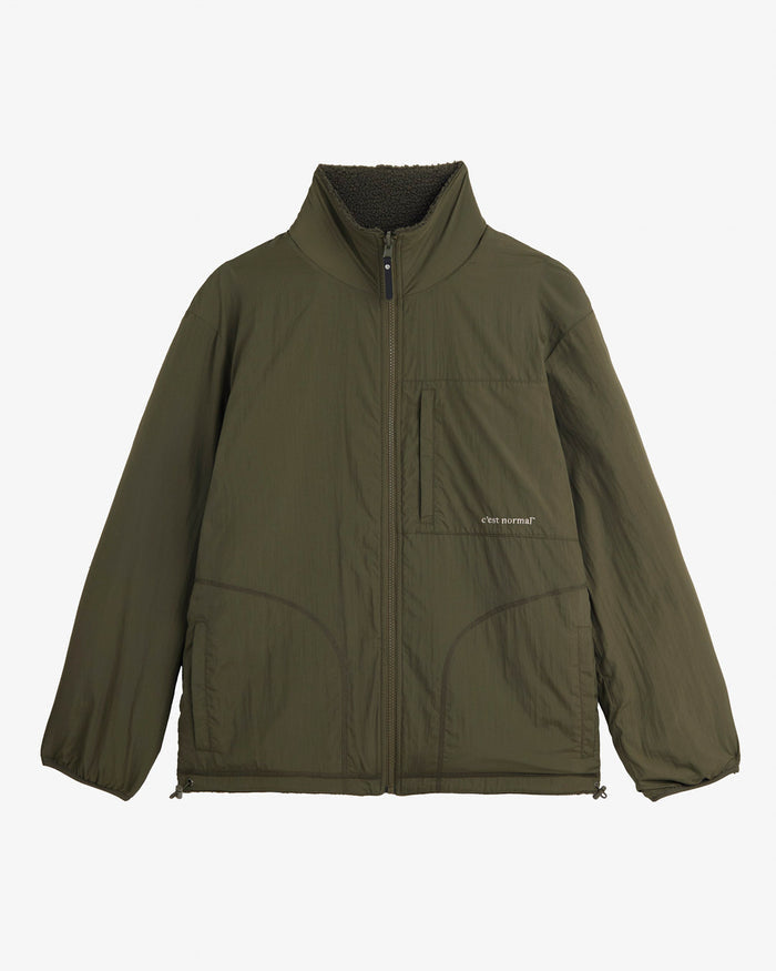 A front view of The Reversible Pile Jacket in Forest Green, laid flat on a white background. The image showcases the smooth tech side, featuring a full front zipper, a zippered chest pocket with the c'est normal logo, and two hand pockets.